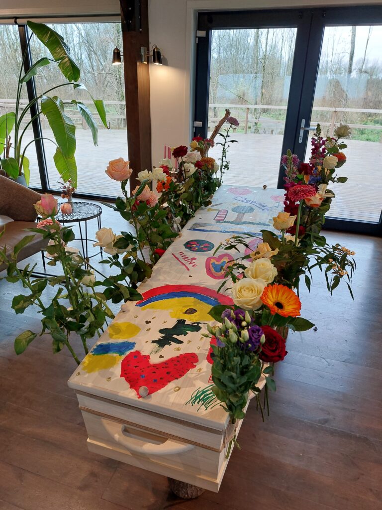 Afscheid op Natuurbegraafplaats Zomerlanden met kist en bloemenband. Met kinderen die kist hebben beschilderd.